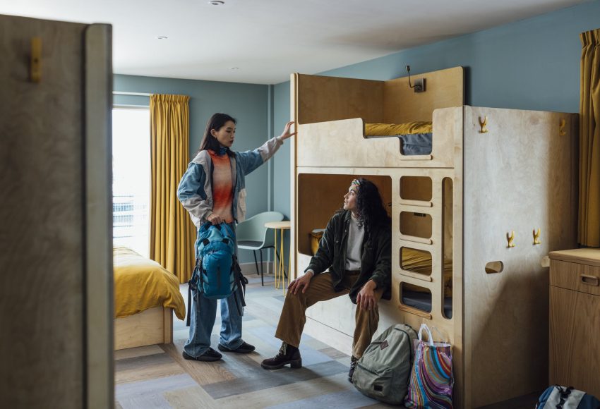 A wide shot of two women wearing warm, casual clothing in a luxury hostel in the seaside town of Amble, Northumberland. They have checked into a dormitory room with bunkbeds. One of them sits on the bed and the other stands, holding her backpack as they talk.